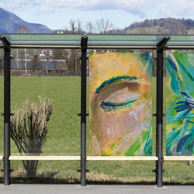 Das Bild zeigt ein Bushaltestelle mit einem gläsernen Wartehäuschen und einem Bildkunstwerk, gemalt auf das Glas der Rückwand.